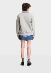 Sudadera gris con hombros caídos, superpuesta a una camisa a rayas azul, combinada con una falda de mezclilla y zapatos negros. Fondo escaso y neutro.