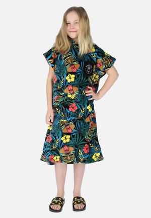 Ung pige med langt blondt hår iført en farverig kjole med tropisk mønster og sorte sandaler, poserer med én hånd på hoften.