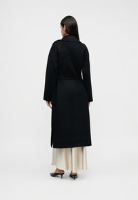 Μαύρο μακρύ παλτό από μάλλινο ύφασμα, με ζώνη στη μέση, φαρδιά μανίκια και κλασικό γιακά, φορεμένο πάνω από μια μακριά φούστα σε εκρού χρώμα.