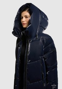 Nafukovací bunda v námořnické modři, vyrobená z lesklého materiálu. Má velkou kapuci, kapsy na zip a prošívaný vzor se čtvercovými sekcemi.