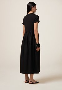 Schwarzes Maxikleid mit kurzen Ärmeln, figurbetontem Oberteil und ausgestelltem Rock. Getragen mit einem schwarzen gefransten Armband und schwarzen Ballettschuhen mit rosa Akzenten.