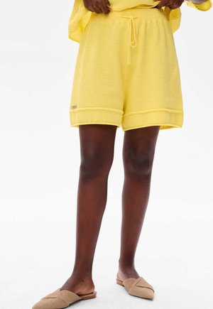 Yellow woven shorts with an elastic waistband, drawstring detail, and rolled hem. Paired with neutral slip-on shoes.