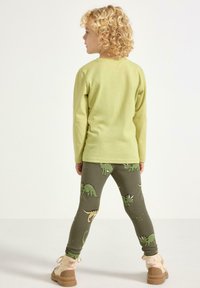 Jasnozielona koszula z długim rękawem i okrągłym dekoltem, połączona z ciemnozielonymi legginsami z wzorami dinozaurów oraz beżowymi butami z pomponami.