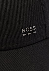 Fekete textil sapka ezüst színű, fémes "BOSS" logóbetűkkel és két díszítő, vízszintes csíkkal a szöveg alatt az elülső panelen.