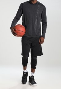 Czarna sportowa bluza z kapturem z logo, w połączeniu z czarnymi siateczkowymi szortami, legginsami kompresyjnymi i czarnymi trampkami, trzymająca piłkę do koszykówki.