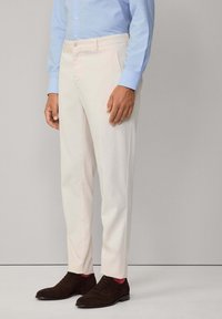 Pantaloni su misura di colore chiaro realizzati in un tessuto liscio. Presentano tasche laterali, una piega discreta e un taglio dritto, abbinati a scarpe marroni scure.