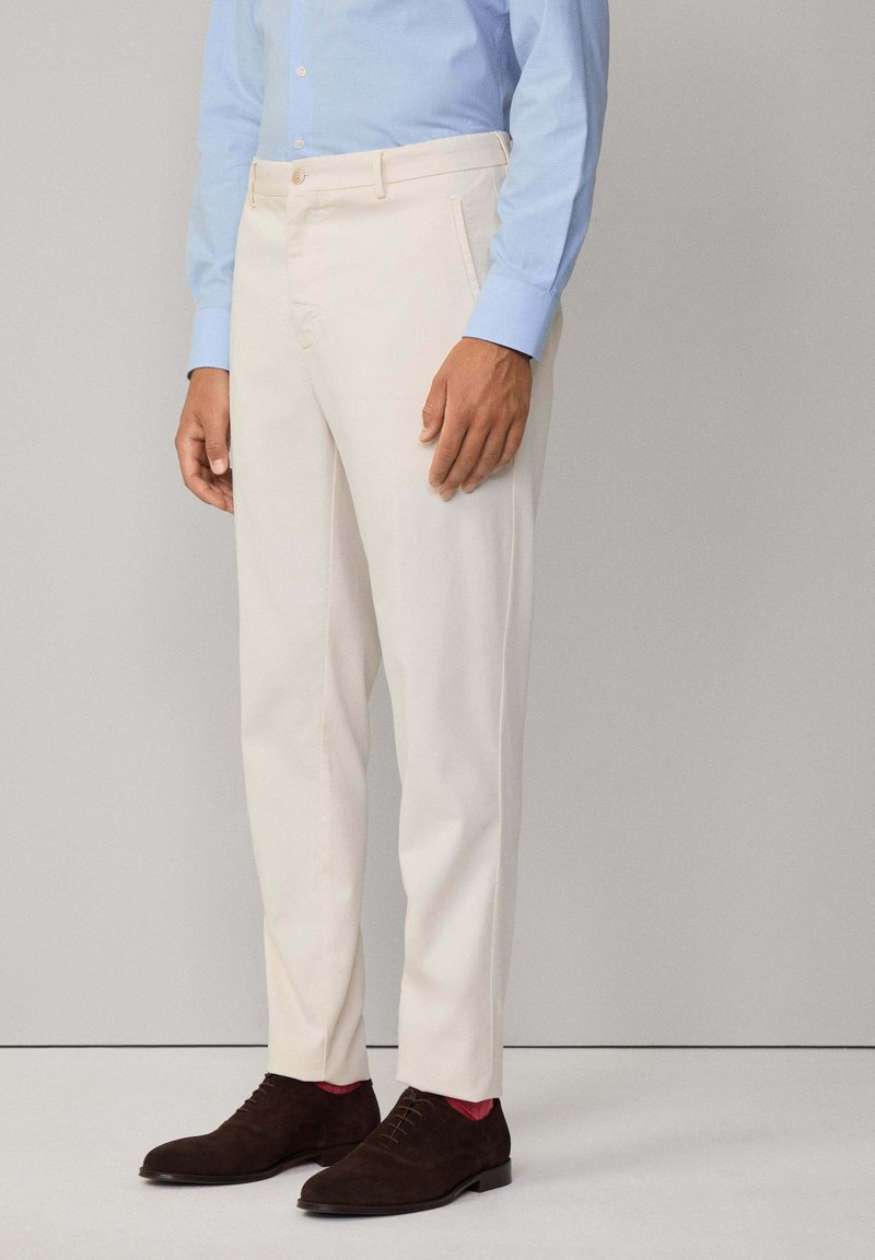 Pantaloni su misura di colore chiaro realizzati in un tessuto liscio. Presentano tasche laterali, una piega discreta e un taglio dritto, abbinati a scarpe marroni scure.