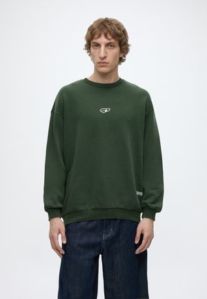 Ung mand med krøllet hår iført en mørkegrøn sweatshirt med et lille hvidt logo og mørkeblå jeans, stående mod en ensfarvet baggrund.