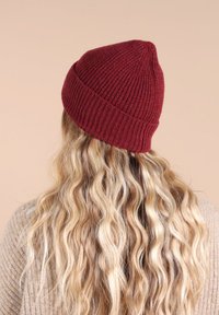 Un bonnet tricoté bordeaux avec une texture côtelée, un dessus effilé et un ajustement flexible, porté sur des cheveux blonds ondulés.