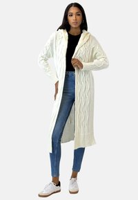 Lunga cardigan in maglia color crema con motivi a treccia, cappuccio e polsini a coste. Indossata aperta su una maglia nera e jeans blu. Presenta tasche.