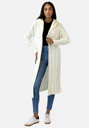 Lunga cardigan in maglia color crema con motivi a treccia, cappuccio e polsini a coste. Indossata aperta su una maglia nera e jeans blu. Presenta tasche.