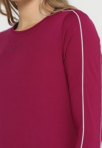 Långärmad skjorta i magenta polyester, med vita sidostripes och en liten svart logotyp på bröstet. Slät textur.