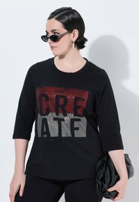 Černé tričko s kulatým výstřihem a rukávy do tří čtvrtin, s flitrovou grafikou v červené, stříbrné a černé barvě. Doplňuje ho psaníčko.