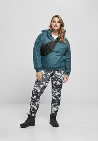 Tirkizna pufer jakna s kapuco, uparen s crno-belim donjim delom od legginga sa tie-dye uzorkom i crnim čizmama na vezanje. Na sebi nosi crnu torbicu preko tela.