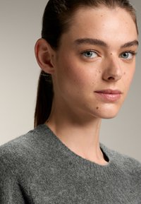 Šedý vlněný svetr s kulatým výstřihem, který má jemnou texturu. Model má světlou pleť, pihy a modré oči, viděný v detailním záběru.
