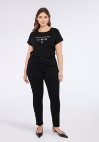 Mujer con cabello castaño hasta los hombros, vistiendo una camiseta gráfica negra, pantalones negros de cintura alta y tacones negros con tachuelas, de pie contra un fondo blanco.