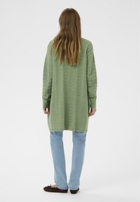 Lange, loszittende trui in lichtgroen met een getextureerd zigzagpatroon, gecombineerd met lichtblauwe jeans en donkerbruine schoenen.