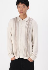 Beige cardigan with a collared neckline, featuring vertical ribbed patterns in a darker shade, and a button-front design. Soft texture.
