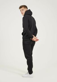 Haut à capuche noir avec une capuche courbée, associé à un pantalon assorti. Coupe slim, texture côtelée, baskets blanches. Mains croisées derrière le dos.