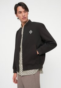 Musternaha musta bomber-jakk, millel on tõusnud krae, embroiditud teemantlogo, ees taskud ja sile kuma viimistlus.