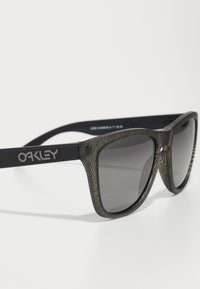 Ochelari de soare negri, cu un cadru texturat, având un model în undă și lentile gri. Logo-ul Oakley este gravat pe un braț.