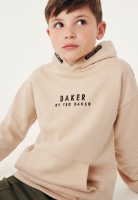 Béžová mikina s kapucí, se přední kapsou, černým logem "BAKER by TED BAKER" a stahovací kapucí s branded akcenty. Měkká textilie.
