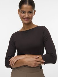 Ung kvinne med pent samlet hår, iført gullringøreringer, en mørk genser med lange ermer og beige bukser, smilende med kryssede armer.