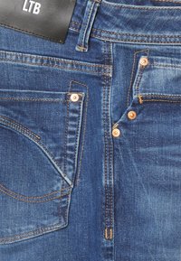 Los jeans de denim azul oscuro cuentan con múltiples bolsillos, remaches de color cobre y costuras de contraste a lo largo de las costuras. Etiqueta de la marca visible en la cintura.
