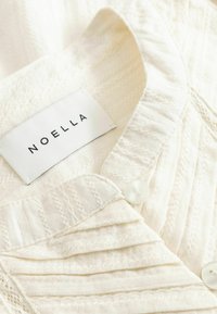 Chemise en tissu blanc texturé avec des motifs délicats, des boutons nacrés et un col, affichant une étiquette de la marque Noella à l'intérieur de l'encolure.