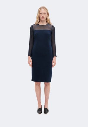 Femme portant une robe bleu marine longueur genou avec manches longues transparentes et empiècement, associée à des mocassins noirs à bout pointu, debout sur un fond uni.