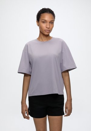 Γυναίκα που φορά φαρδιά ανοιχτογκρι κοντομάνικη μπλούζα και μαύρα σορτς, στέκεται μπροστά σε απλό λευκό φόντο.