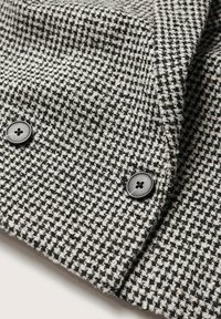 Tissu pied-de-poule noir et blanc avec boutons noirs croisés sur la manche texturée d’un manteau en laine.