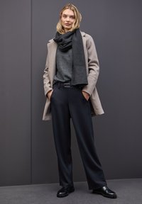 Mujer de pie contra una pared oscura, vistiendo un abrigo beige, una bufanda gris oscuro, un suéter gris, pantalones negros y zapatos negros, con las manos en los bolsillos.