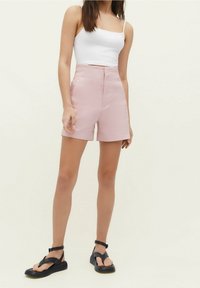 Pantalones cortos ajustados de color rosa claro con una cintura alta y bolsillos frontales, combinados con una camiseta sin mangas blanca ajustada y sandalias planas negras.