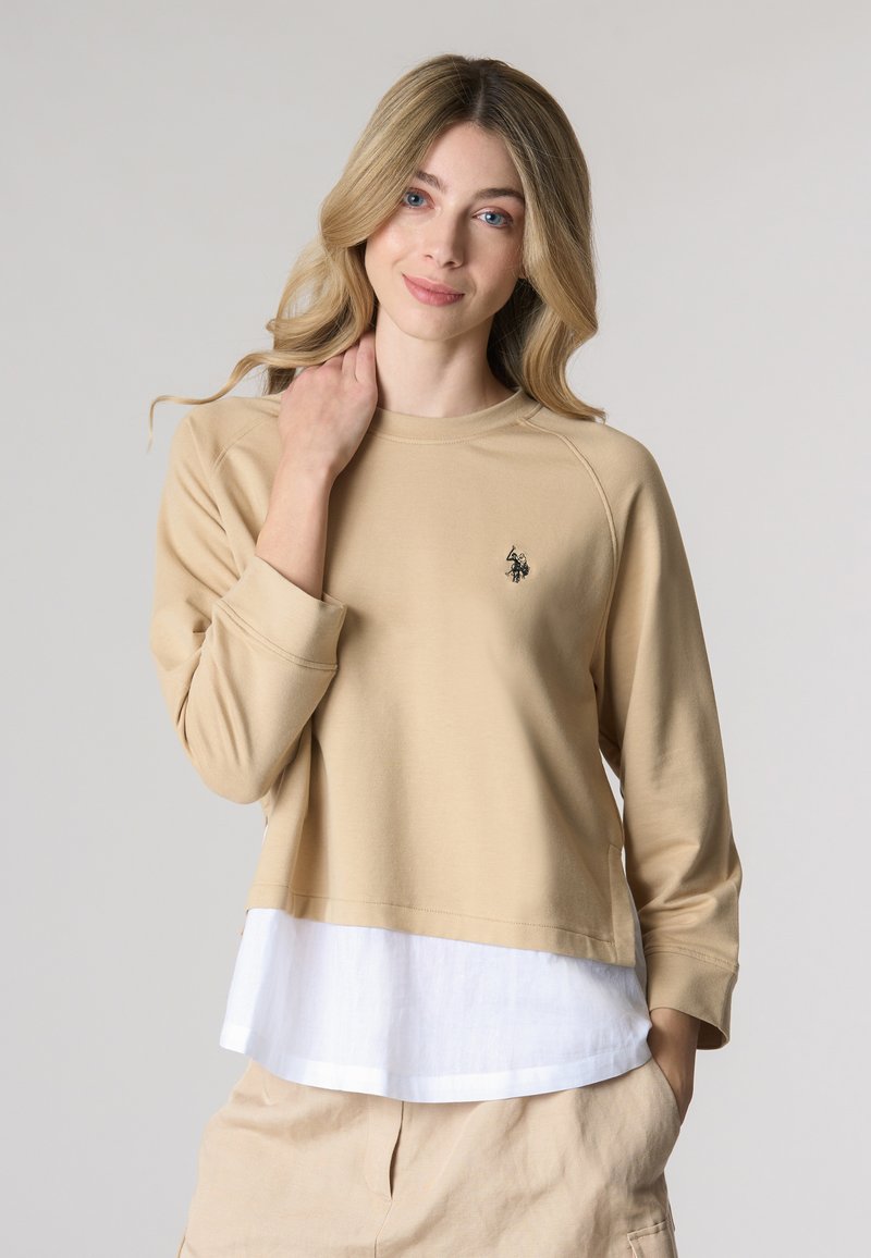 Felpa beige con maniche 3/4, caratterizzata da un orlo a strati bianco e un piccolo emblema nero sul petto. Texture liscia, design casual.