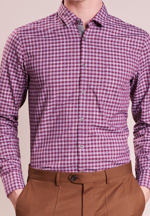 Mężczyzna w koszuli na guziki z długim rękawem w fioletowo-białą kratę, włożonej do brązowych spodni, ręce w kieszeniach.