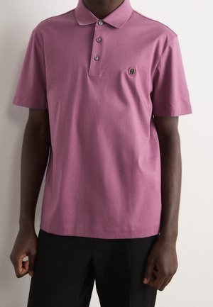 Man draagt een paars poloshirt met korte mouwen, drie knopen en een klein logo op de linkerborst, gecombineerd met een zwarte broek.