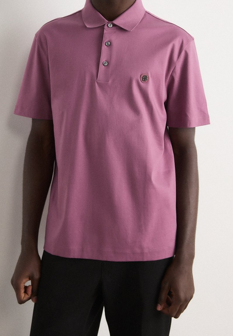 Homme portant un polo violet à manches courtes avec trois boutons et un petit logo sur la poitrine gauche, associé à un pantalon noir.