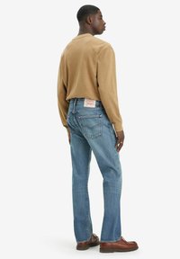 Lichtbruin overhemd met lange mouwen, gecombineerd met lichtblauwe jeans. De jeans hebben een klassieke pasvorm, vijf zakken en een merklabel achterop.