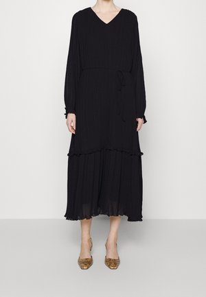 Femme portant une longue robe noire plissée avec des manches longues et un col en V, associée à des talons pointus marron à motif peau de serpent.