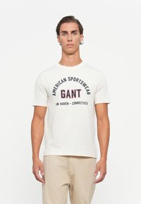 Mann trägt ein weißes Kurzarm-T-Shirt mit dem Aufdruck "American Sportswear GANT New Haven - Connecticut", kombiniert mit beigen Hosen, im Stehen vor einem weißen Hintergrund.