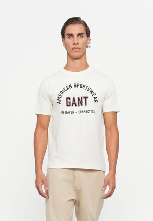 Mies, joka on pukeutunut valkoiseen lyhythihaiseen T-paitaan, jossa lukee "American Sportswear GANT New Haven - Connecticut", yhdistettynä beigearvoisiin housuihin, seisoo valkoista taustaa vasten.