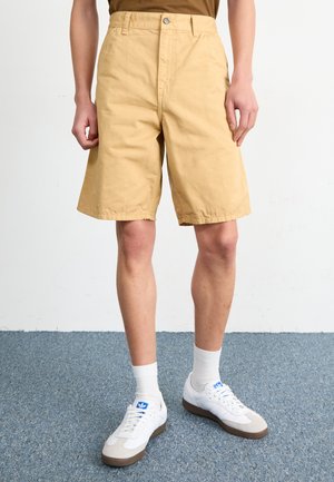 Bermudas de algodón beige con corte recto, con cierre de botón y trabillas para cinturón. Combinadas con zapatillas blancas y calcetines.
