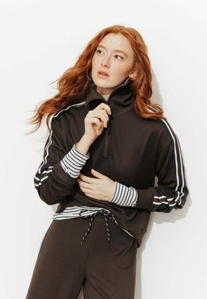 Femme aux cheveux roux et taches de rousseur portant un haut marron à fermeture éclair avec des manches rayées et un pantalon assorti, ajustant la fermeture éclair en regardant sur le côté.