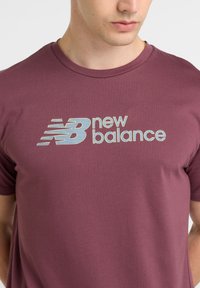 Burgo raudonas medvilnės marškinėliai su mėlyna ir balta dryžuota "New Balance" logotipu. Trumpomis rankovėmis ir apvaliu kaklu, maloni tekstūra.