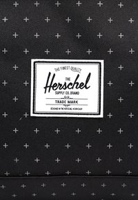 Tissu noir avec un motif de petits signes plus blancs et une étiquette rectangulaire blanche de la marque Herschel Supply Co. au centre.