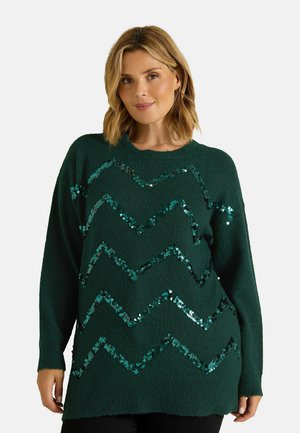 Femme aux cheveux blonds mi-longs portant un pull vert foncé avec des motifs en sequins brillants en zigzag sur fond clair.