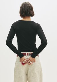 Top a maniche lunghe nera abbinato a pantaloni chiari con un motivo testurizzato e una cintura rossa. Gli accessori includono più anelli.