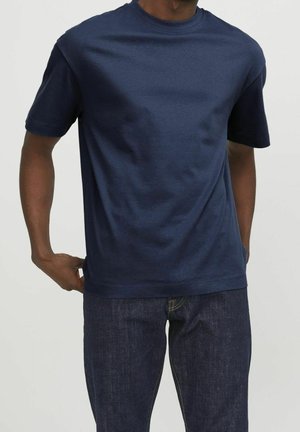 Hombre con camiseta azul marino lisa de manga corta y vaqueros azul oscuro, de pie frente a un fondo claro y liso.