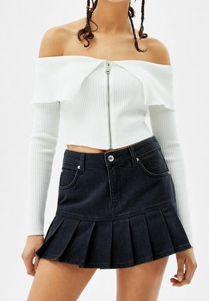 Vrouw draagt witte off-shoulder geribbelde top met rits en zwarte geplooide denim minirok, laat de buik en armen langs het lichaam zien.